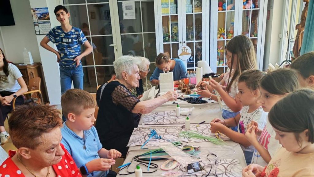 Group of children and seniors sit around a long craft table, making bead and ribbon decorations together in a bright room.