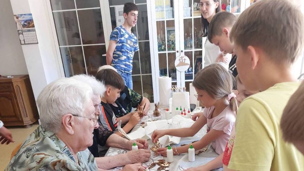 Old man and children crafting together at a table with glue and wood pieces in a bright room.