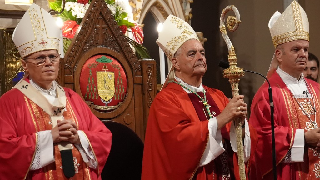 Three Catholic bishops in red vestments and white miters stand during a church ceremony; the central bishop holds a gilded crozier beside an ornate carved throne with flowers behind them.