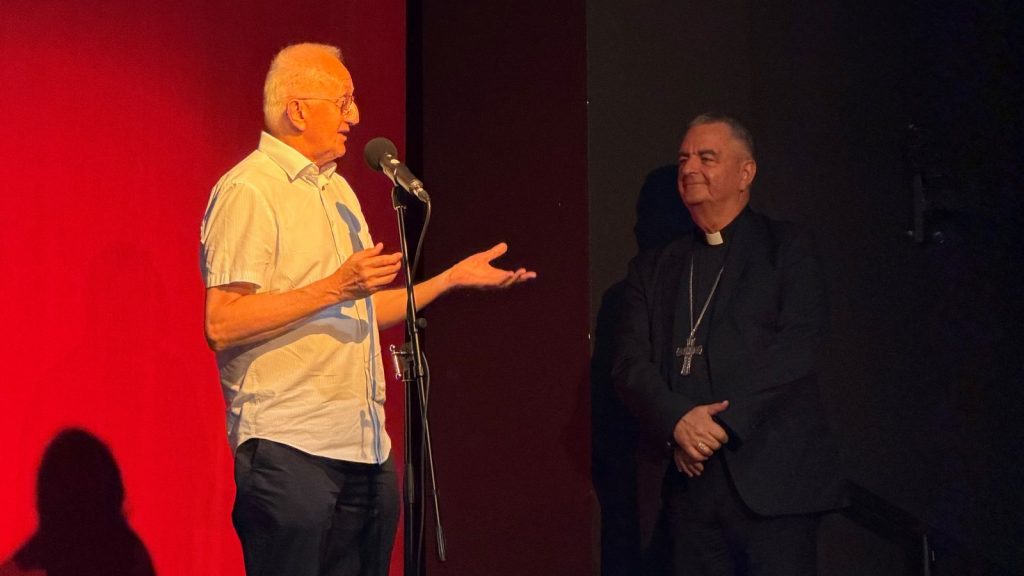 Two men on a stage: one in a light shirt speaking into a microphone, gesturing with his hands, while a clergyman in black robes and a cross watches.