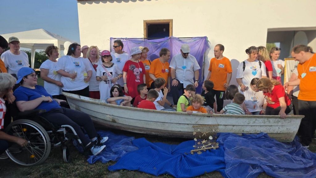 Group of adults and kids gathered around an old rowboat on a blue tarp during an outdoor event; a person in a wheelchair sits at left watching the scene.
