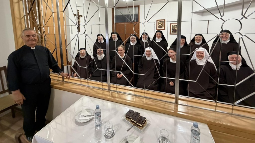 Priest stands at a counter beside a decorative metal grate, smiling at a group of nuns in habits behind it; table with water bottles and small cakes in front.