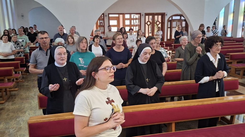 Congregation standing in prayer inside a church with red pews and arched windows, including two nuns in black habits near the foreground.