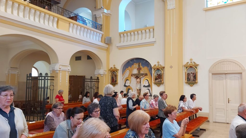 Parishioners sit on wooden pews inside a sunlit church with arches and religious murals on the altar wall.