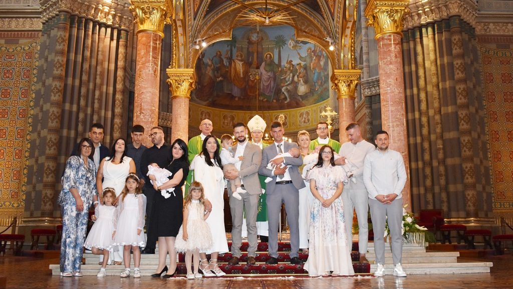 Group of adults and children in formal attire posing on church steps, with clergy and a bishop in the ornate sanctuary behind them during a ceremony.