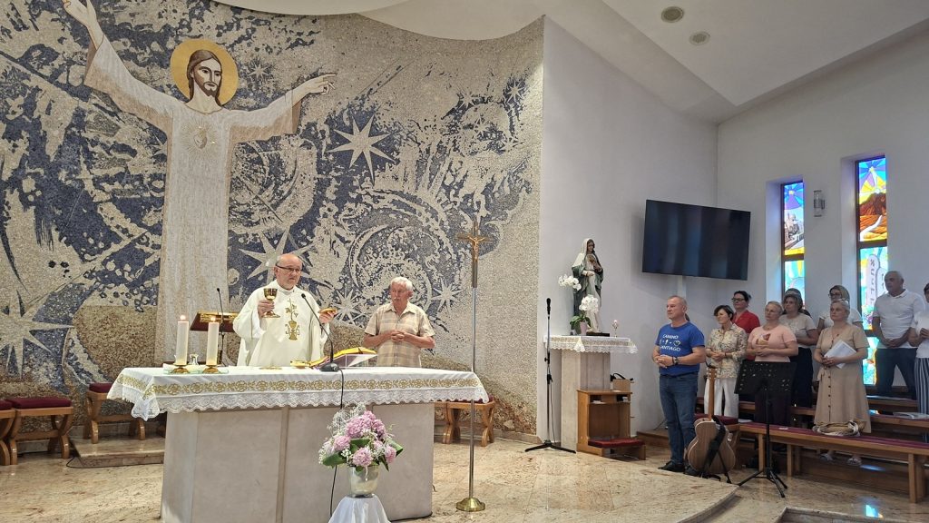 Priest at the altar during Mass holding a chalice and host, with a man standing beside him and a large mosaic of Jesus behind.