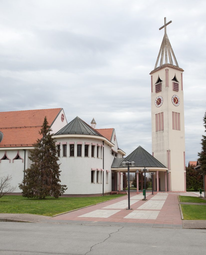 White church complex with pink trim and a tall clock tower topped by a cross, along a paved walkway under cloudy sky.