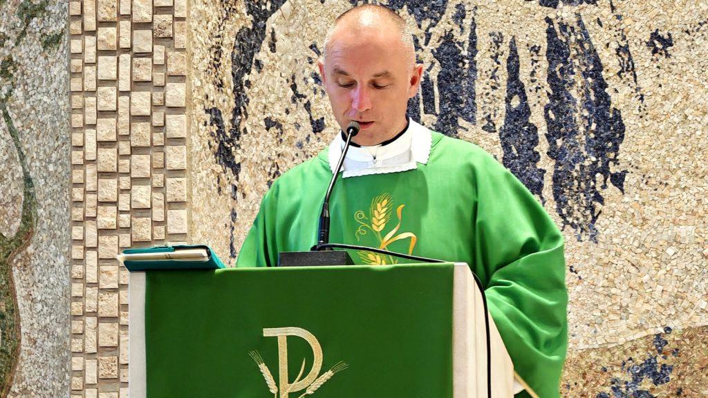 Priest in green vestments delivering a sermon at a lectern with a mosaic backdrop.