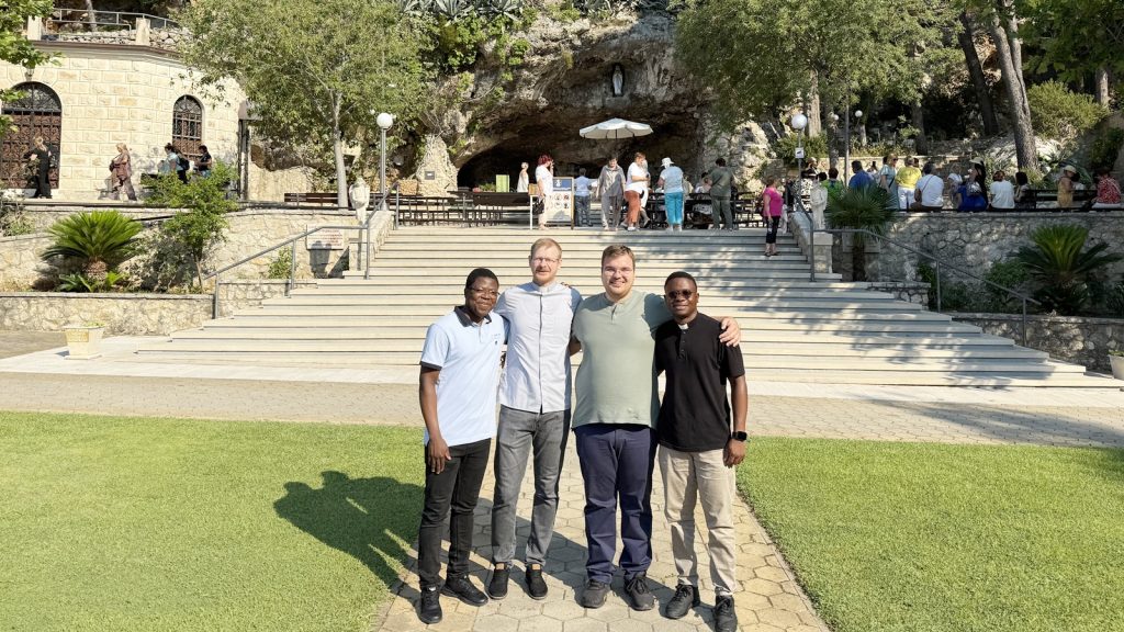 Four men stand arm-in-arm on a paved path in front of stone stairs at a park, posing for a group photo with a sunny courtyard in the background.