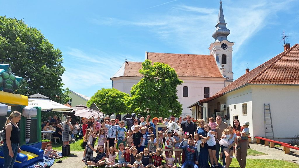 A large group of children and adults posing for a photo outdoors at a sunny community event, with a church and trees in the background and tents on the left.