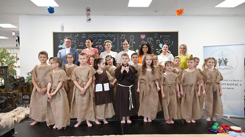 Group of children in burlap costumes posing with teachers in a classroom stage setting, barefoot, with a chalkboard backdrop and seasonal decorations behind them.