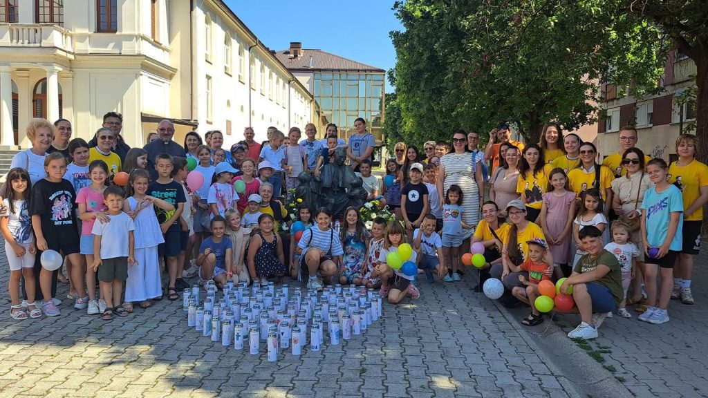 Large group of children and adults posing outdoors in front of a building, with colorful balloons and a row of water bottles arranged on the ground in front of them.