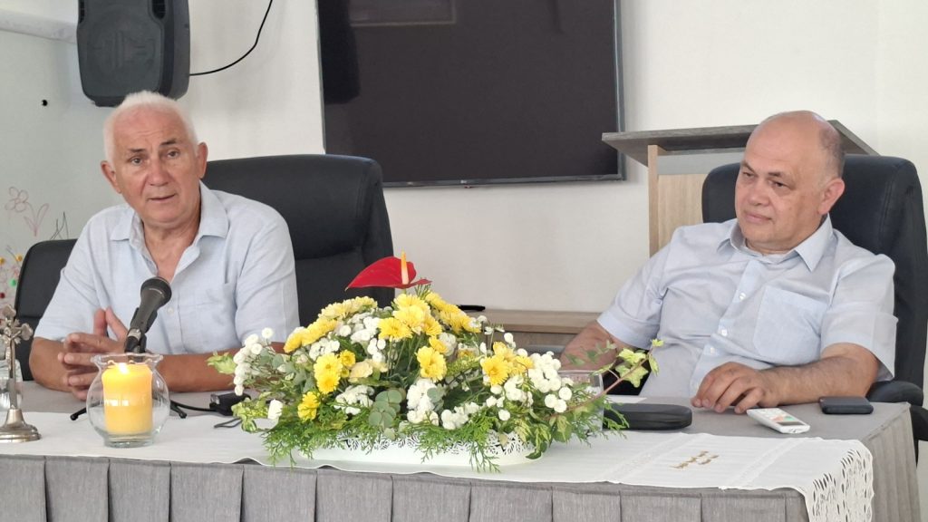 Two men sit at a conference table; one speaks into a microphone with a candle and floral centerpiece between them in a formal setting.