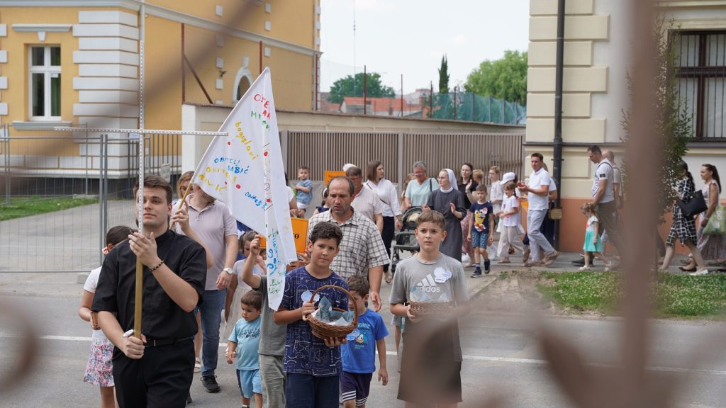 Group of children and adults walking in a street parade, carrying a large banner and baskets, led by a man in black clothes.