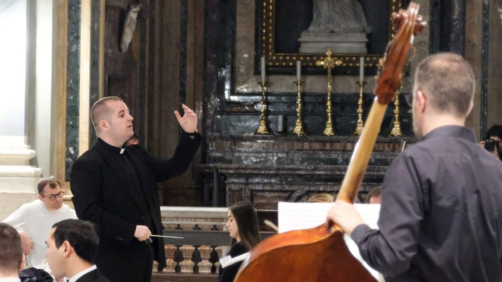 Conductor in a black suit raises his hand while leading musicians inside a church with a marble altar in the background and a large string instrument in the foreground.