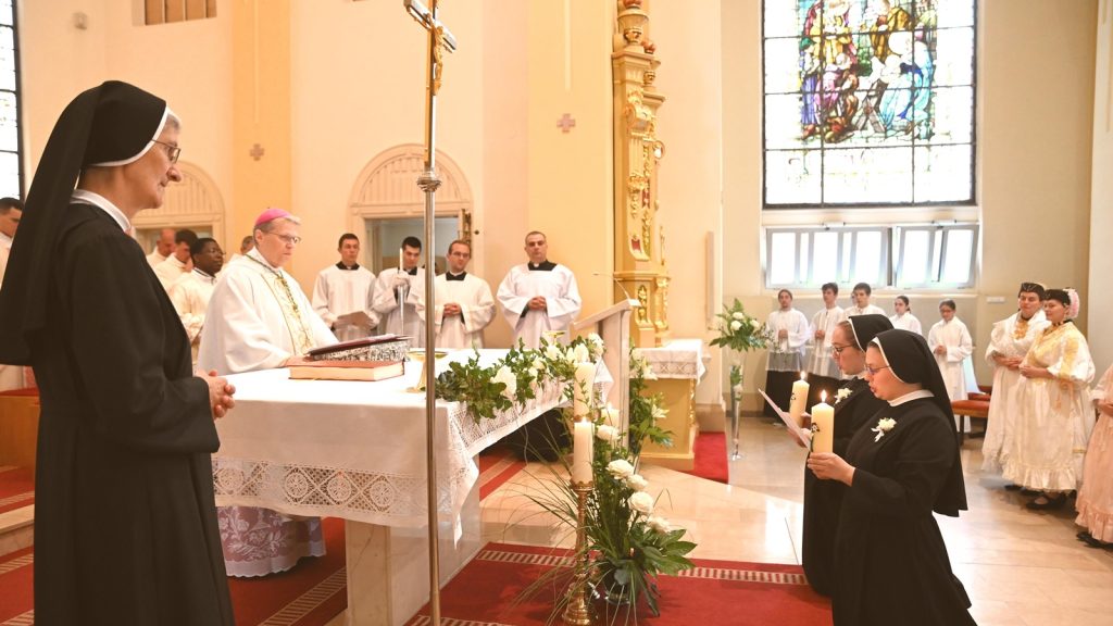 Nun in a black habit stands at left as a bishop in white liturgical vestments leads at the altar, with flowers and candles nearby.