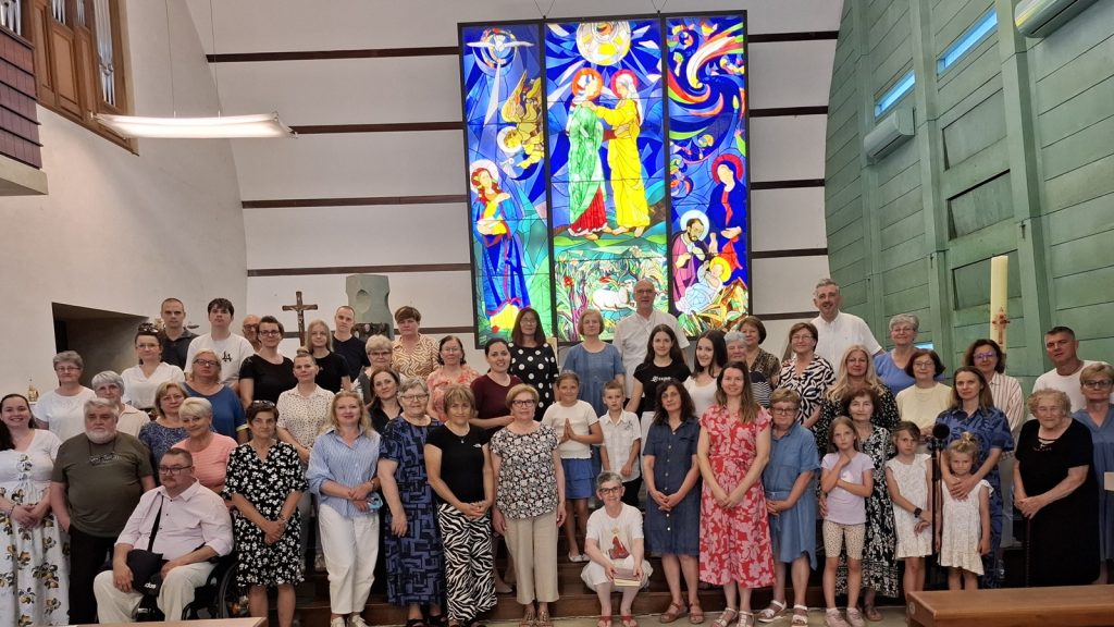 Group of parishioners of various ages posing in a church, with a large colorful stained-glass window behind them and a cross nearby.