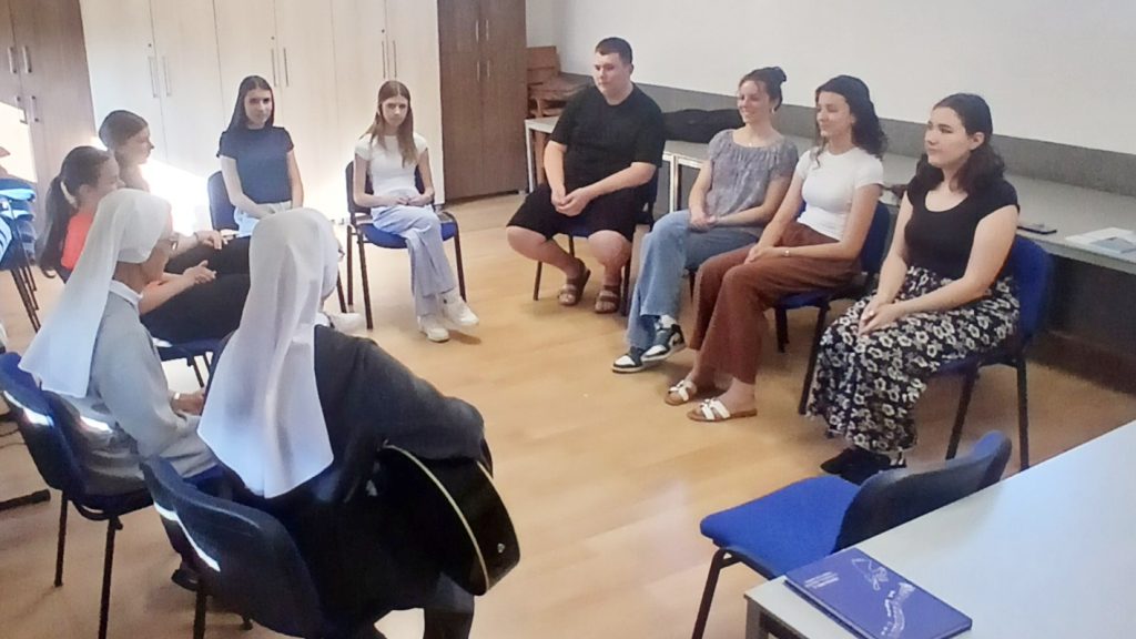 Group of people sitting in a circle in a bright room, including two nuns in white veils, engaged in a discussion.