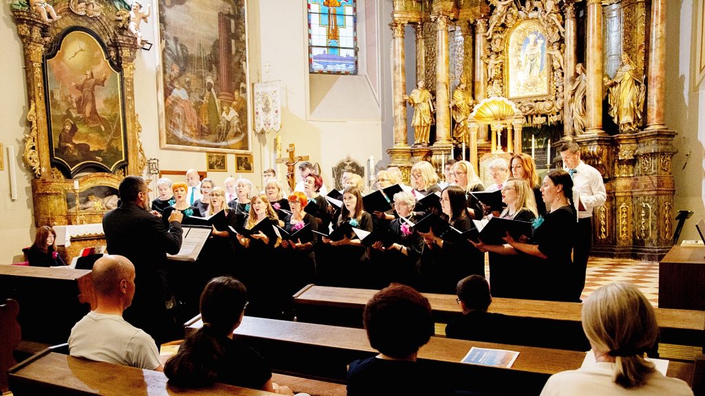 A choir of men and women in black robes sings under a conductor in a gilded church interior with ornate altarpiece and stained glass behind them.