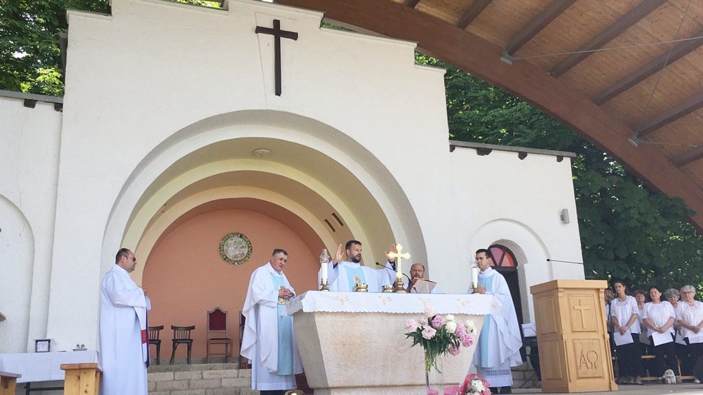 Outdoor Catholic Mass at a white church altar with priests in white vestments around a stone altar and a cross centerpiece.