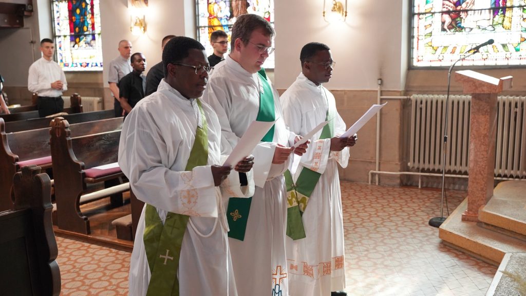 Three priests in white robes with green stoles read from papers during a church service, with stained glass windows in the background.