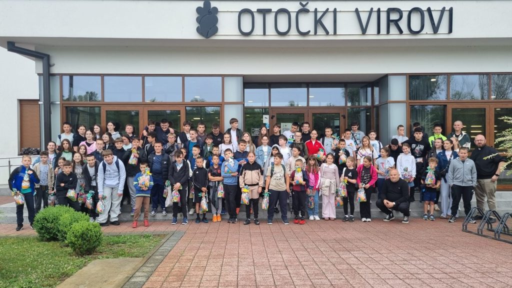 Group of students (and a few adults) posing outside a building with the 'OTOČKI VIROVI' sign, each holding gift bags.