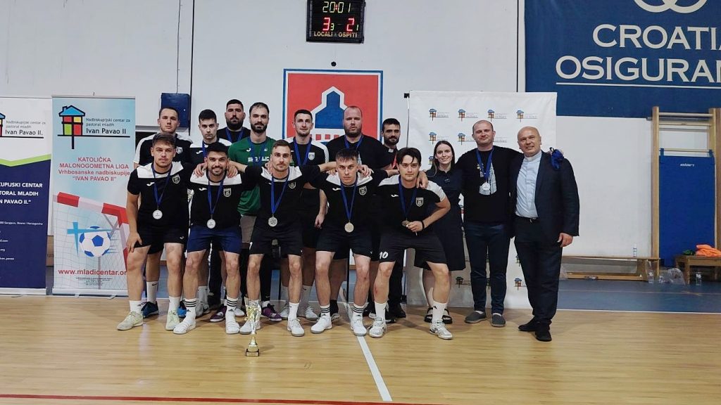 A group of athletes in black uniforms posing with medals around their necks and a trophy on the floor in front, indoors after a game celebration.
