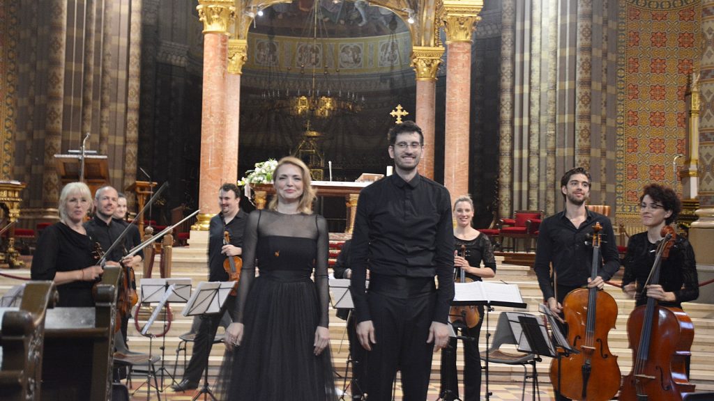 Musicians in black attire pose on a church altar stage with violins and cellos, ornate columns in the background and a gold cross above them
