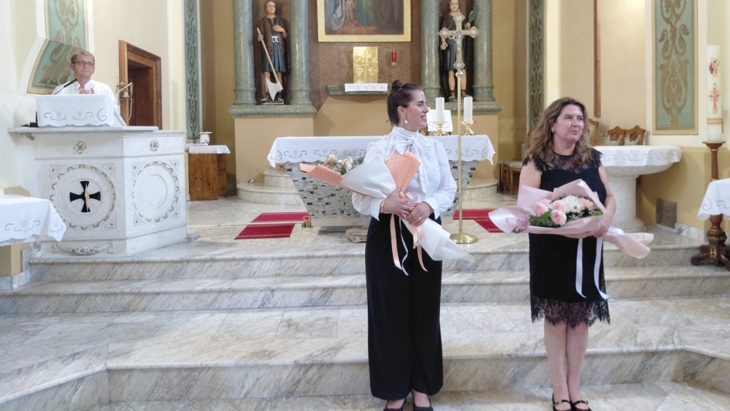 A man speaks from a marble pulpit in a church while two women in front hold pink bouquets, standing on marble steps near the altar.