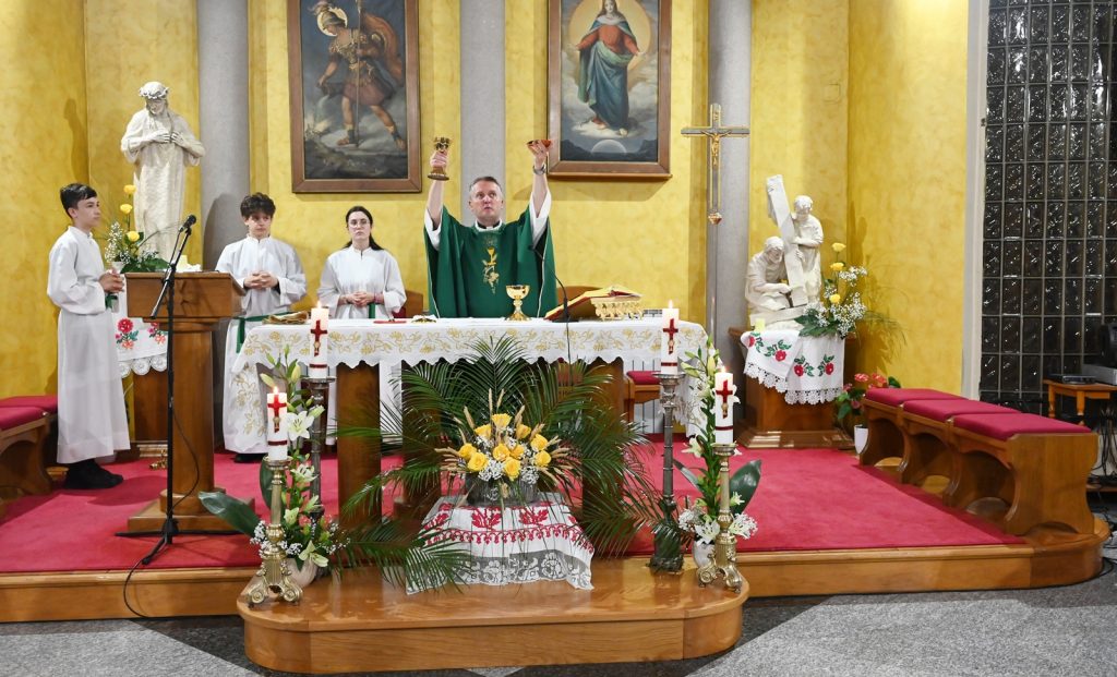 Priest in green vestments raises chalice and host at the altar during Mass, with three servers in white robes nearby.