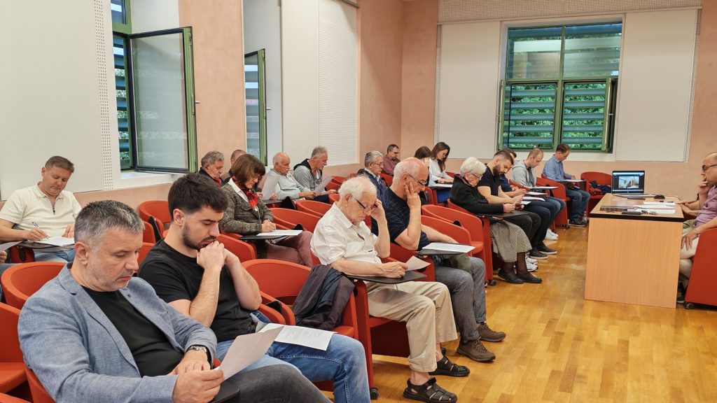 Audience of adults seated in orange chairs, taking notes during a meeting in a classroom-like auditorium.