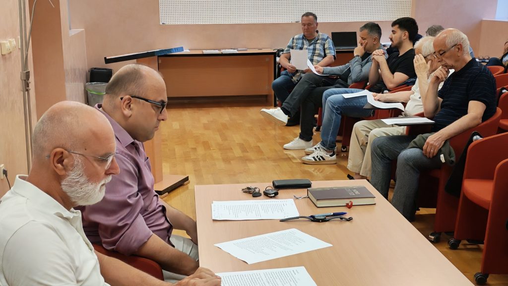 Two men sit at a table at the front of a room with papers and a book, while a group of people watch and review documents in a small meeting or class setting.