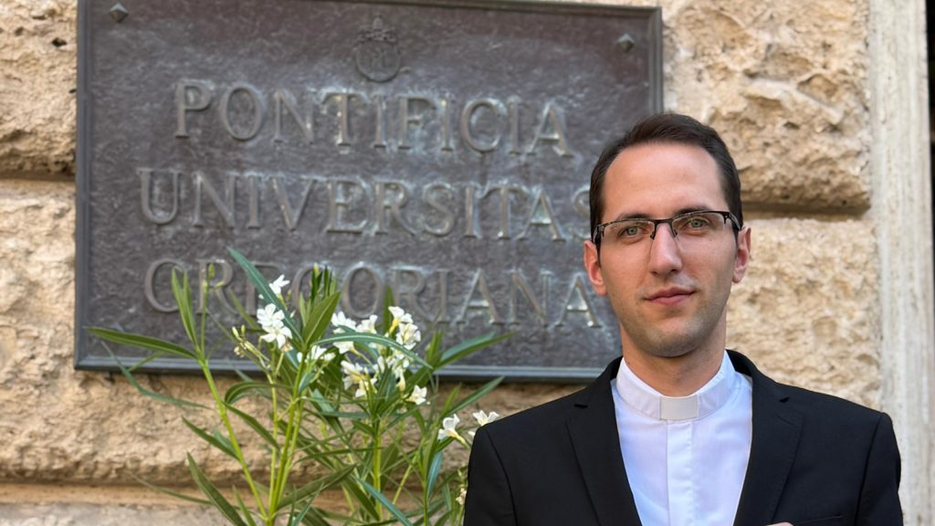 Clerical-collar man in front of a stone wall and plaque reading Pontificia Universit&agrave; Gregoriana, with white flowers nearby.
