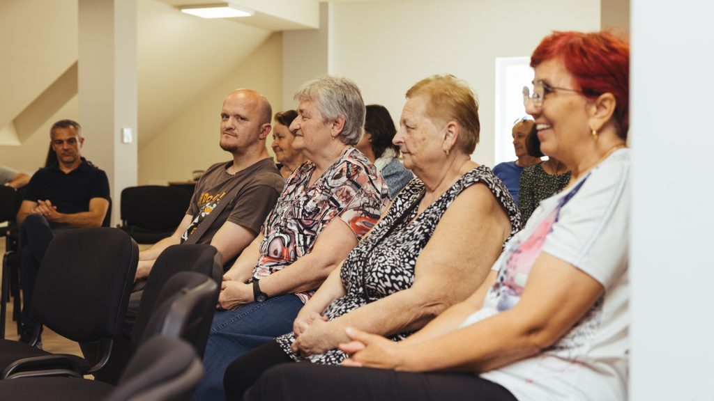 Mixed-age adults seated in a conference room, listening attentively.