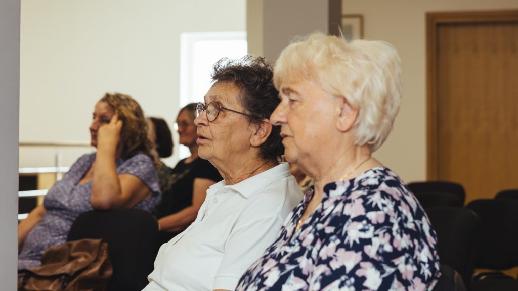 Older adults seated in a row of chairs in a meeting room, listening attentively to a speaker.