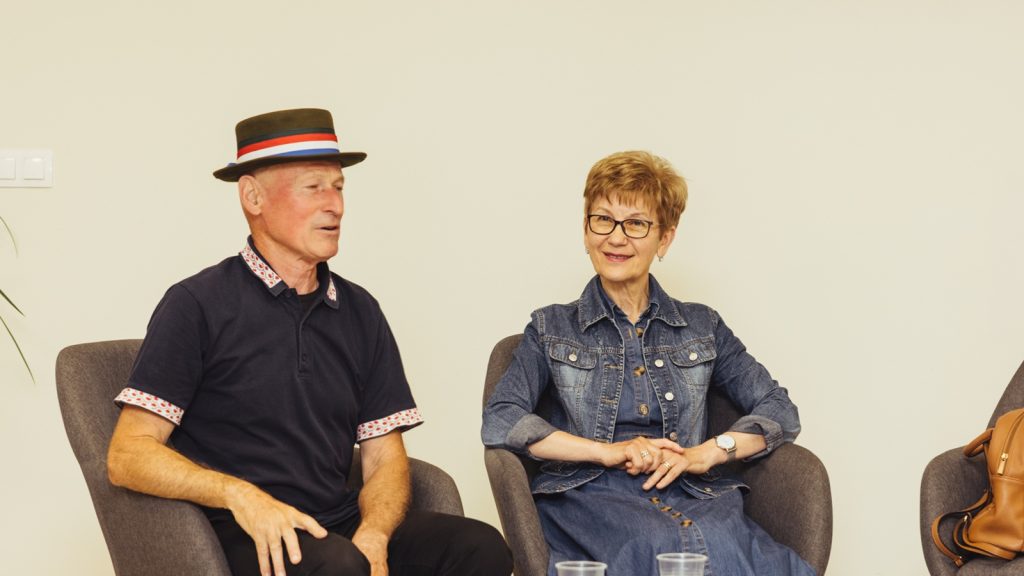 Two older adults sit in chairs and chat in a bright room; man wears a brown hat with red, white, and blue band and a navy polo, woman in a denim jacket and glasses.
