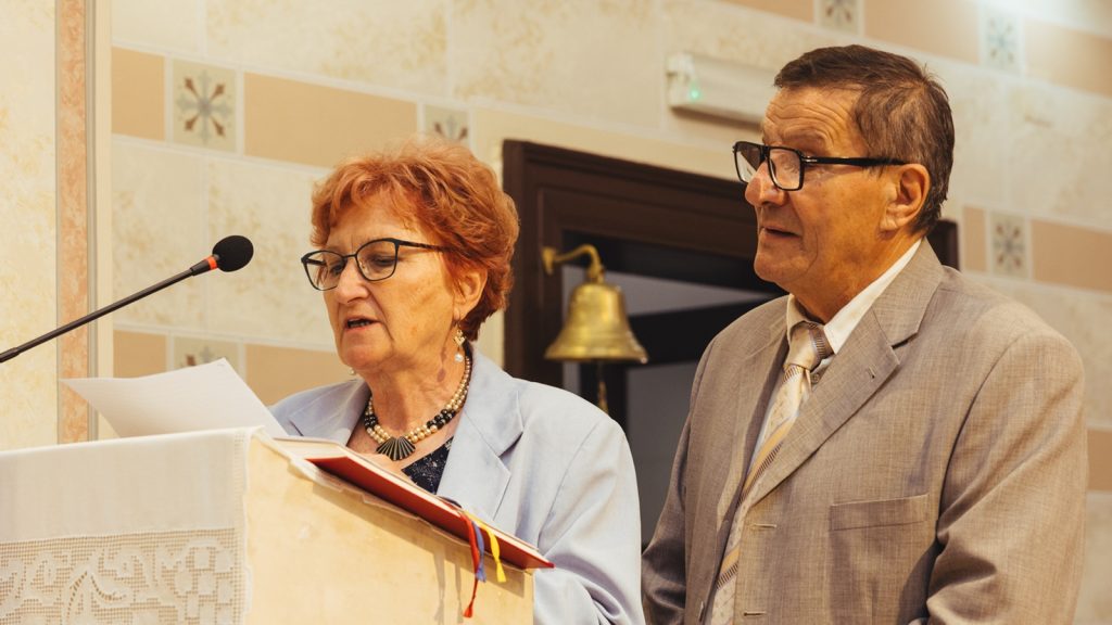 Elderly woman at a lectern reading from papers, microphone in front; man in a suit stands beside her.