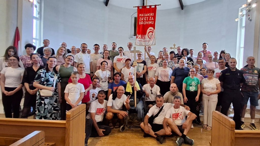 Large diverse group posing for a group photo in a church hall, with a red banner and a cross behind them, all facing the camera.