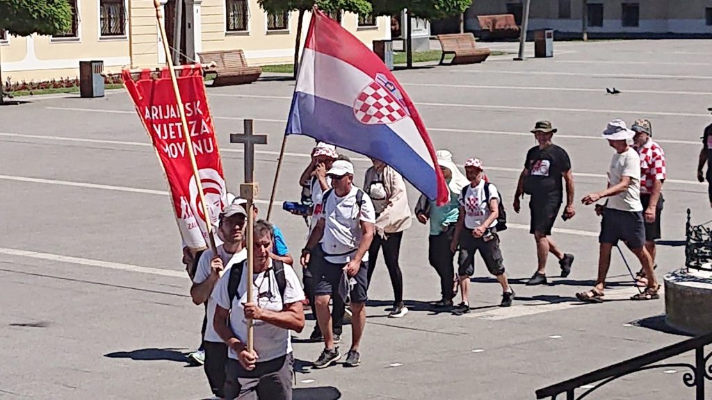 Group of people walking in a parking lot, carrying a Croatian flag and a red banner with a crest/inscriptions.