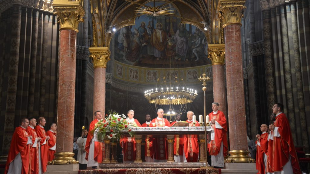 Catholic priests in red vestments celebrate Mass at a gilded marble altar in a grand, ornate cathedral interior with a large chandelier overhead.
