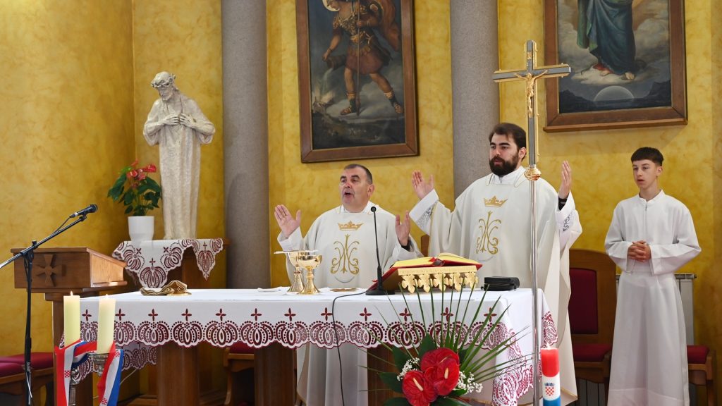 Catholic priests at altar in white vestments celebrate Mass, with open liturgical book and chalice on the altar.