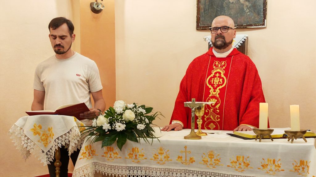 Priest in red ornate vestment stands at the altar with chalice and candles during a church service.