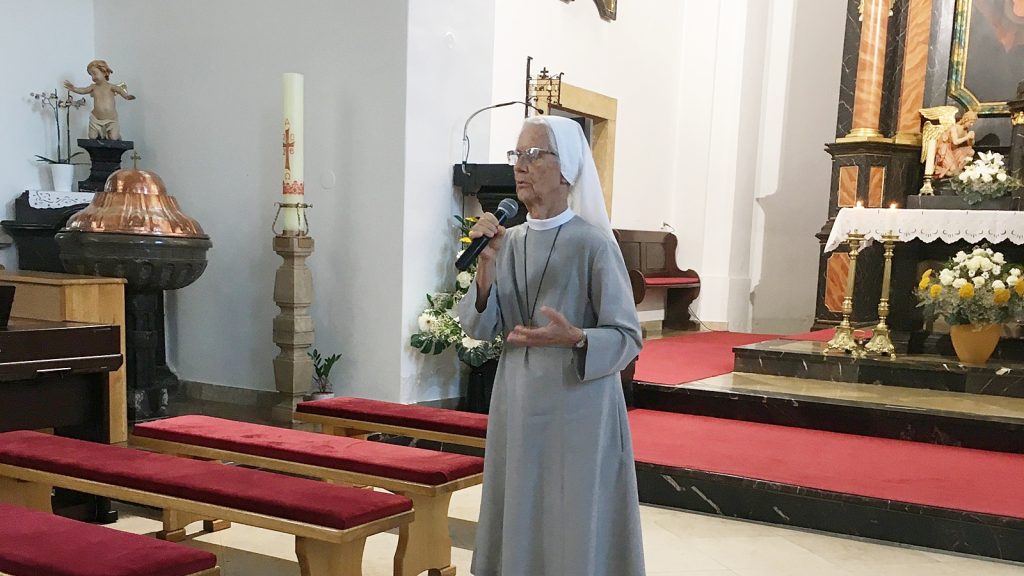 A nun in a light gray habit speaks into a handheld microphone inside a church, near the altar with candles and flowers.