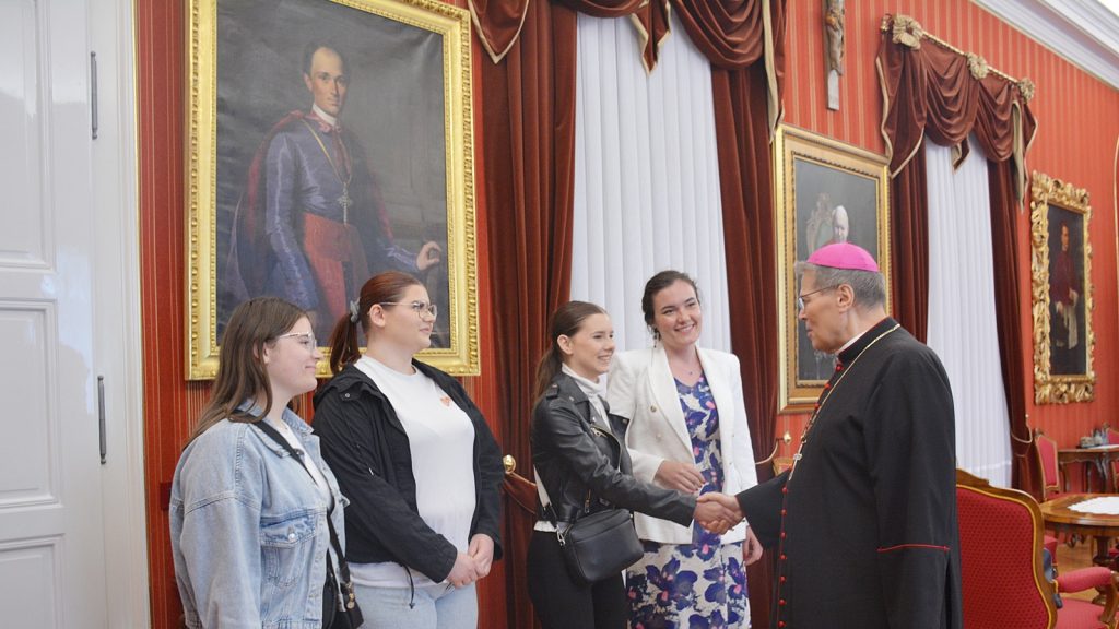 Clergyman in black robes with pink zucchetto shaking hands with four smiling young women in a formal, portrait-filled room.