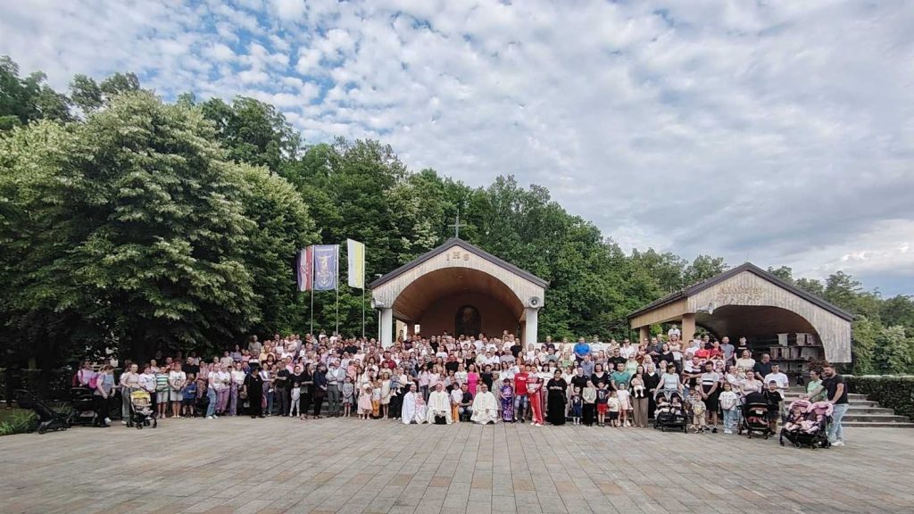 Large group of people posing for a group photo outdoors in front of a small chapel/shrine with a cross, trees in the background, and flags nearby.