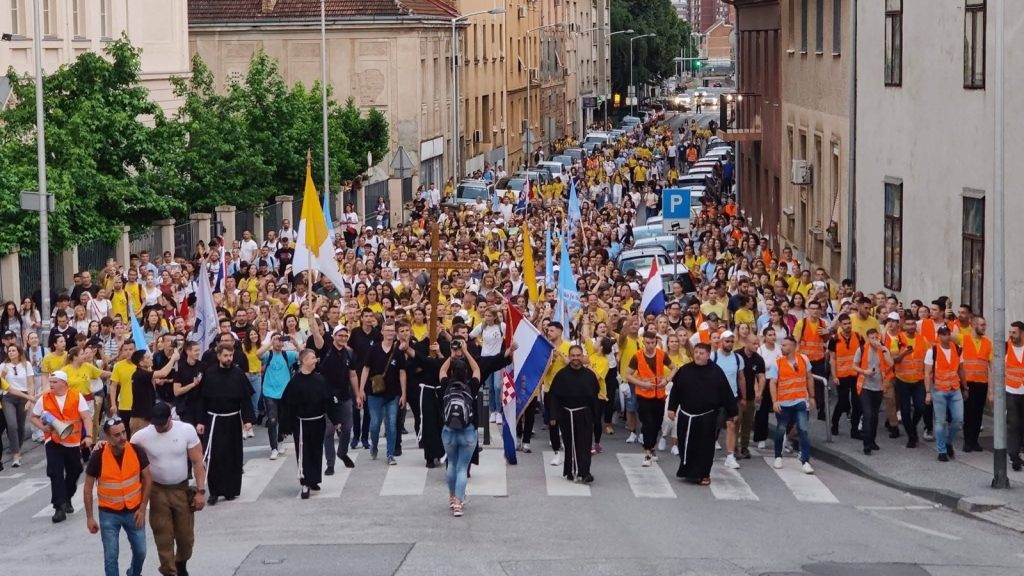 Large crowd marching down a city street in yellow shirts and orange vests, with flags at the front and clergy in black robes leading the way.