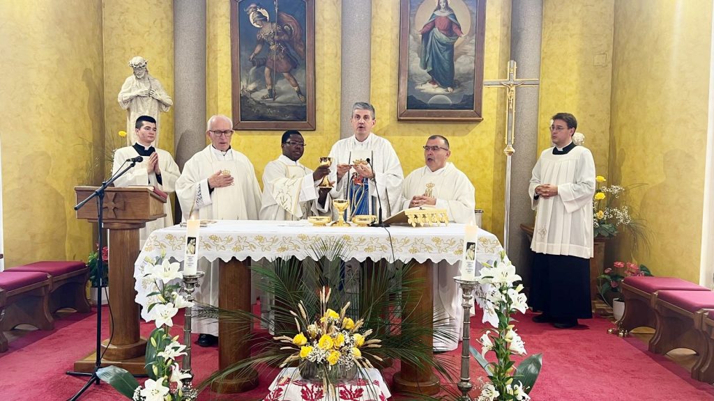 Catholic Mass in progress: priests in white vestments at an ornately decorated altar, raising chalices for blessing with assistants nearby.