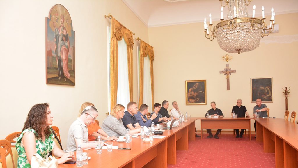 Group of people seated around a long conference table in a formal room with a crystal chandelier and religious artwork on the walls.