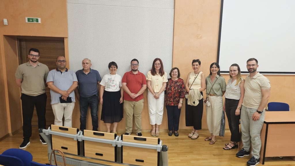 Group of eleven adults standing in a classroom or lecture hall, posing for a photo in front of an orange wall and a projector screen.
