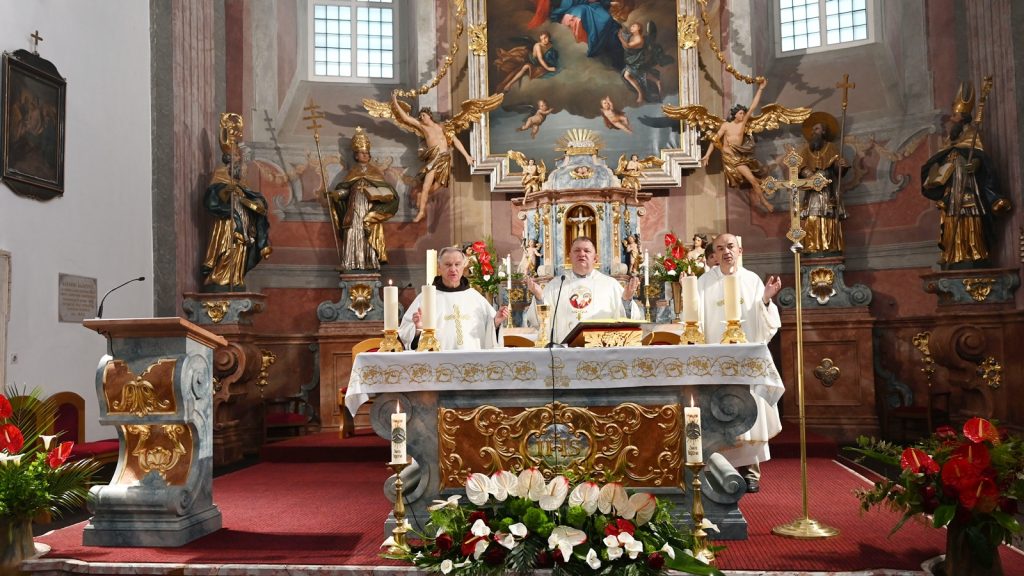 Three priests in white vestments celebrate Mass at an ornately decorated church altar with gold accents, candles, and floral arrangements.
